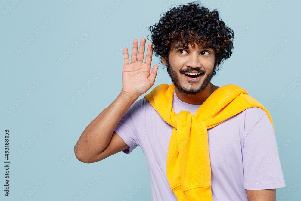 Young bearded Indian man 20s years old wear white t-shirt try to hear you overhear listening intently isolated on plain pastel light blue background studio portrait. People emotions lifestyle concept Young bearded Indian man 20s years old wear white t-shirt try to hear you overhear listening intently isolated on plain pastel light blue background studio portrait. People emotions lifestyle concept