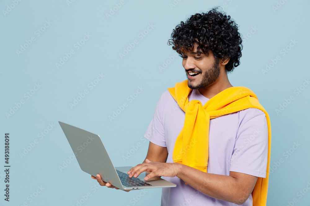 Excited young bearded Indian man 20s years old wears white t-shirt hold use work on laptop pc computer typing browsing chatting send sms isolated on plain pastel light blue background studio portrait. Excited young bearded Indian man 20s years old wears white t-shirt hold use work on laptop pc computer typing browsing chatting send sms isolated on plain pastel light blue background studio portrait.