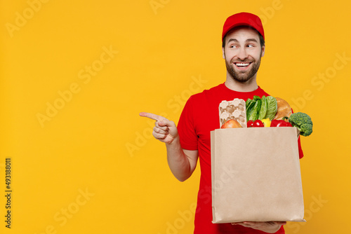 Delivery guy employee man in red cap T-shirt uniform workwear work as dealer courier hold craft paper bag with grocery food point finger aside on workspace area isolated on plain yellow background photo