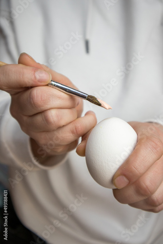 Paint brush in woman's hand and clean white chicken egg. A woman prepares to start painting eggs in Easter holidays in April. DIY Easter preparation concept. People and traditions. Vertical background