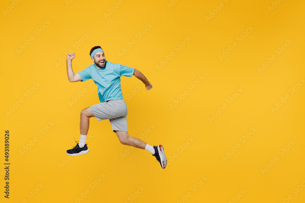 Full body side view strong young fitness trainer instructor sporty man sportsman wear headband blue t-shirt jump high run fast look aside isolated on plain yellow background. Workout sport concept