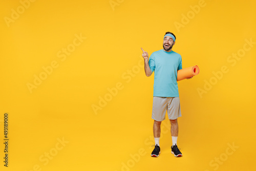 Full body young fitness trainer instructor sporty man sportsman in headband blue t-shirt point index finger overhead workspace hold yoga mat isolated on plain yellow background. Workout sport concept