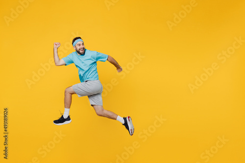 Full body side view strong young fitness trainer instructor sporty man sportsman wear headband blue t-shirt jump high run fast look aside isolated on plain yellow background. Workout sport concept