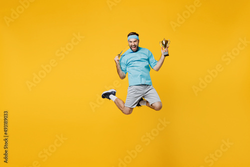 Full body young fitness trainer instructor sporty man sportsman in headband blue t-shirt golded medal hold winer cup jump high isolated on plain yellow background. Workout sport motivation concept photo