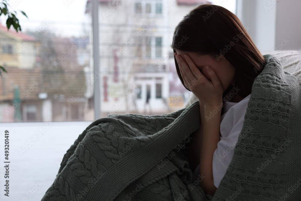 Fototapeta premium Sad young woman wrapped in warm plaid crying at home, space for text. Loneliness concept