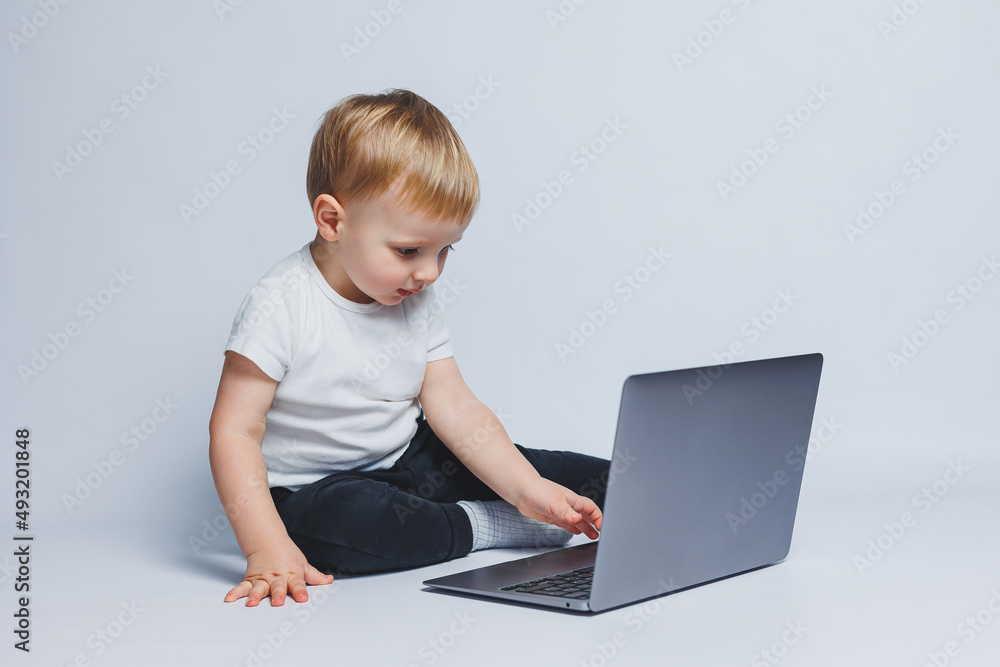 A little boy 3-4 years old sits with a laptop on a white background. A child in a white T-shirt and black trousers looks at a laptop. Modern children A little boy 3-4 years old sits with a laptop on a white background. A child in a white T-shirt and black trousers looks at a laptop. Modern children