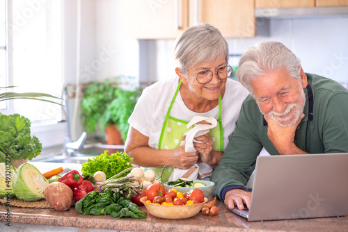 Attractive senior couple preparing vegetables in home kitchen by trying new recipes on the web from laptop. Smiling Caucasian seniors enjoying healthy nutrition
