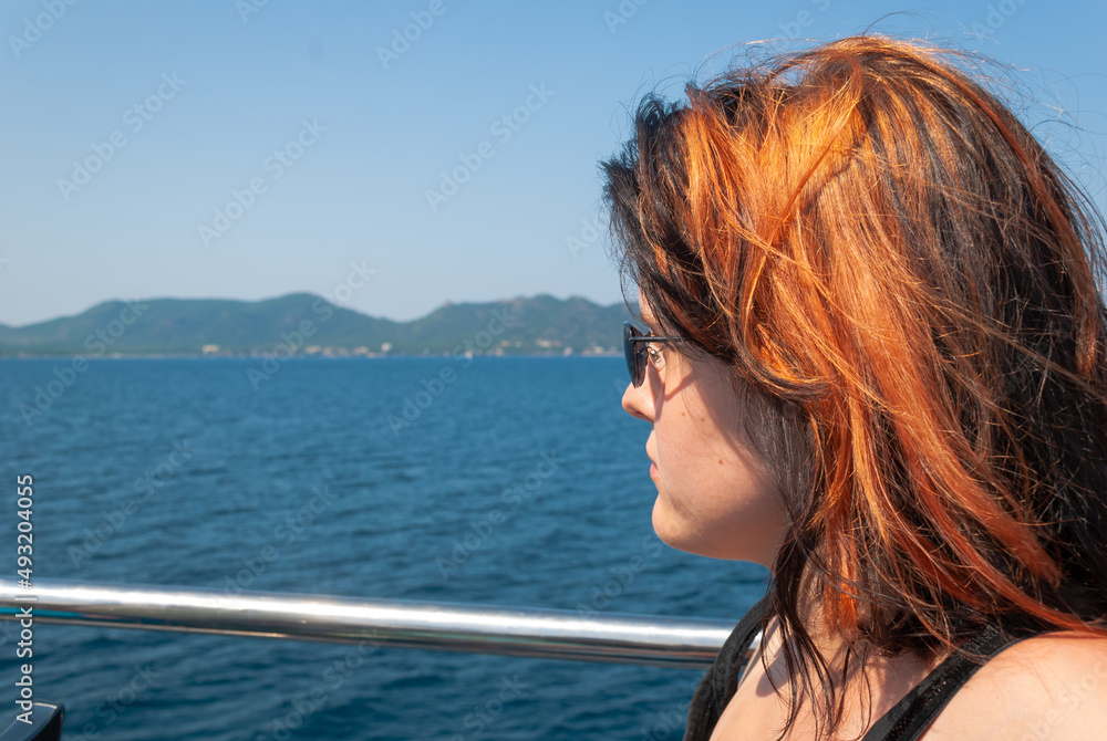 Woman with black orange hair, wearing sunglasses sitting on a ship looking at the mediterranean sea, island in the background, close-up horizontal