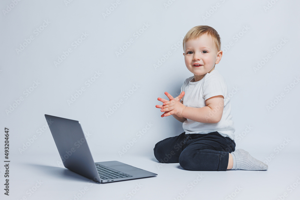 A smart boy 3-4 years old sits with a laptop on a white background. A child in a white T-shirt and black trousers sits at a laptop and looks at the camera. Modern children A smart boy 3-4 years old sits with a laptop on a white background. A child in a white T-shirt and black trousers sits at a laptop and looks at the camera. Modern children