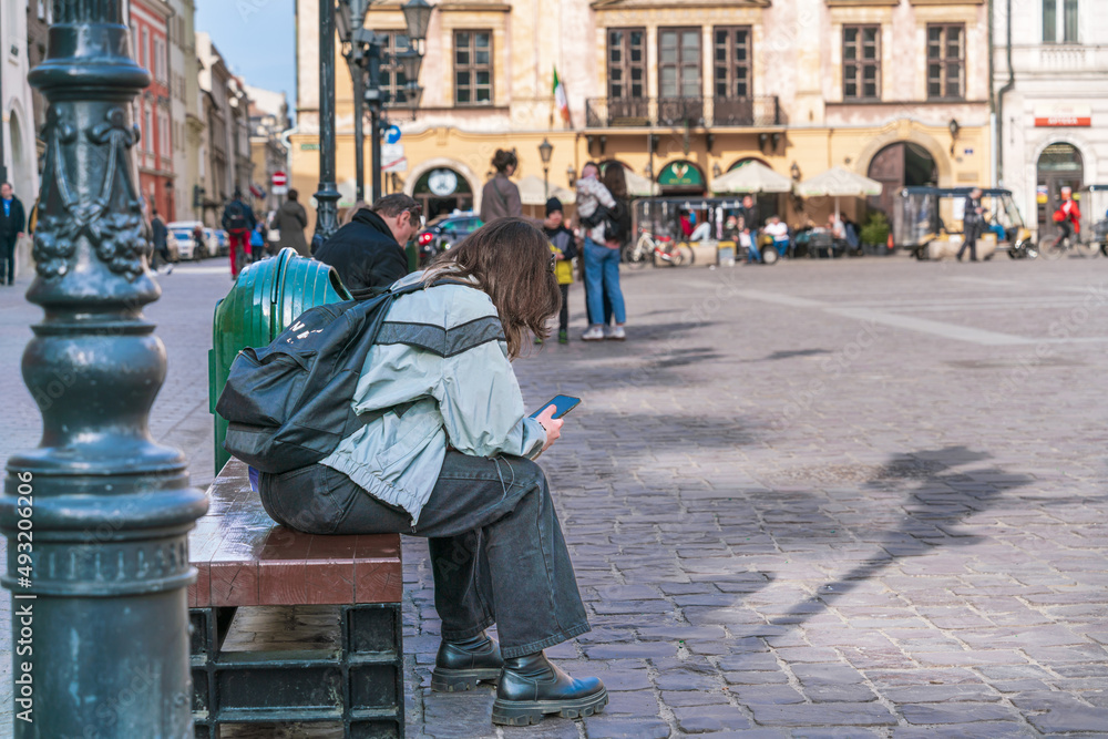 A young girl is sitting on a bench in the city square with a backpack on her back and a smartphone in her hands on a sunny day
