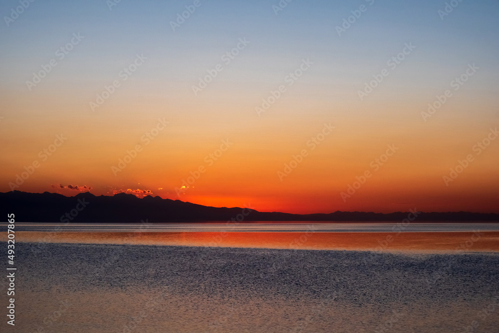 Beautiful sunset on mountain lake with mountain ridge silhouette. Colorful sunrise with orange sky on Issyk-kol lake. Travel, tourism in Kyrgyzstan concept.