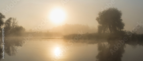 Sunrise over the river. Early foggy morning. Reflection of the sun in the water. Tree by the river. Panorama of several photos.