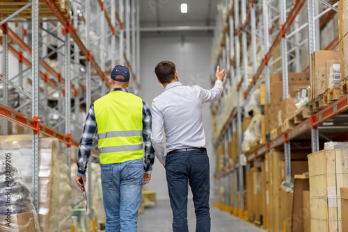 wholesale, logistic business and people concept - manual worker with clipboard and businessman walking along warehouse