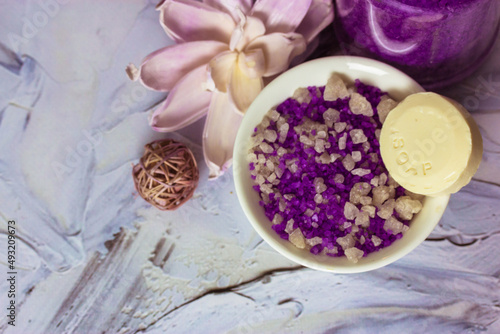 Sea salt and soap. Composition of lilac sea salt in pellets, soap bar, lotus flower on white brushstrokes background flatly. Spa treatments, relaxation, feminine concept. Copy space.