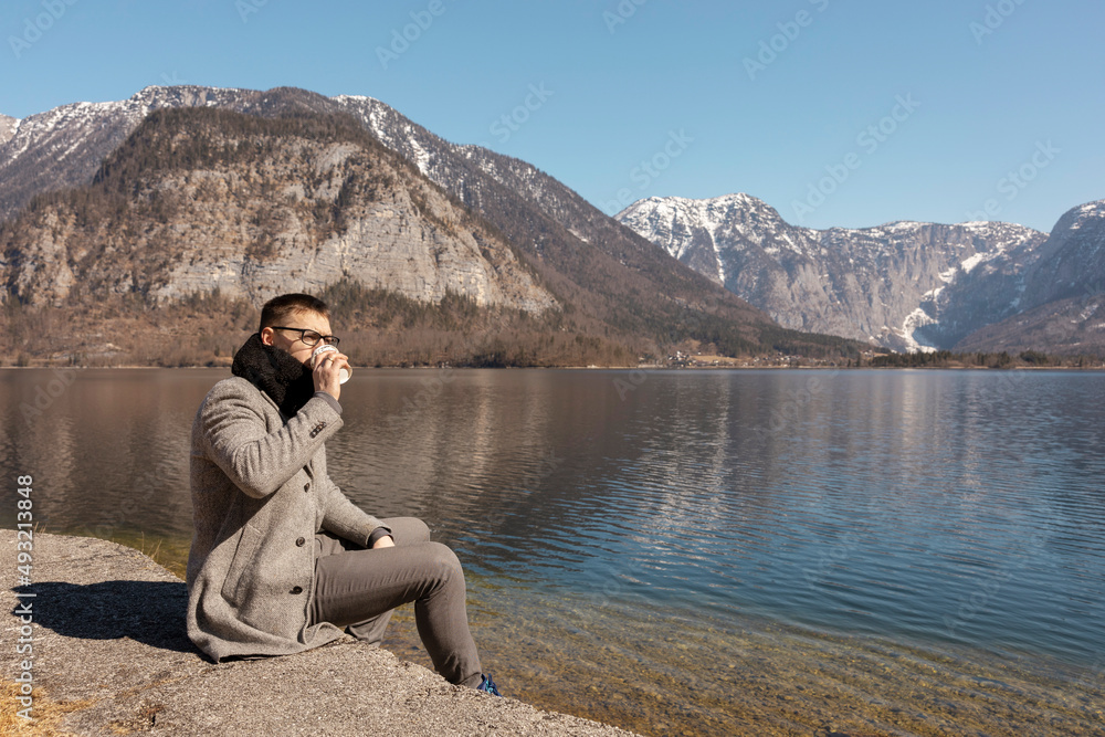 Young adult man sitting outdoors, drinking coffee and enjoying mountains, lake, good weather, blue sky and sun. Beautiful landscape. Time with yourself, dreaming, relaxation, mental health. Holiday.