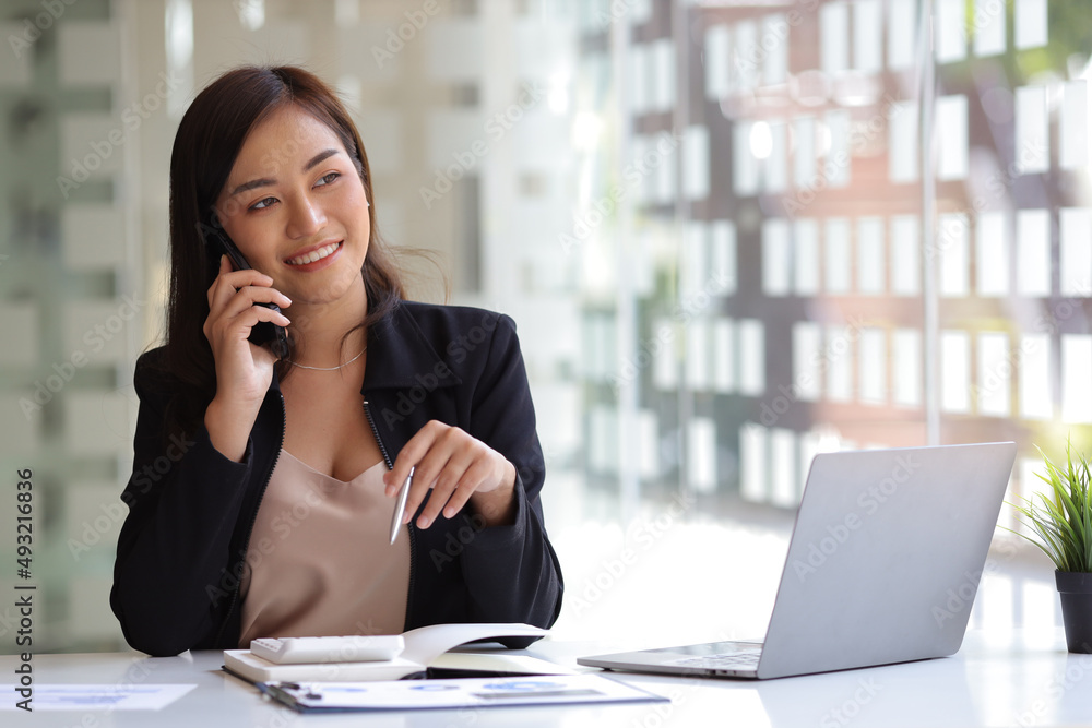 Happy businesswoman sitting at desk with laptop and talking with somebody on her mobile phone while working office. Happy businesswoman sitting at desk with laptop and talking with somebody on her mobile phone while working office.
