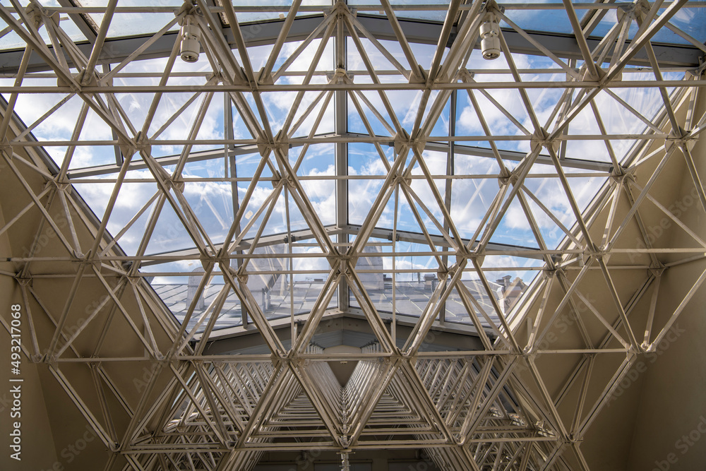 Modern architectural fiction. A metal structure supporting contemporary gallery glass dome. Transparent ceiling letting in sunlight, glass dome blue sky and rooftop in background.