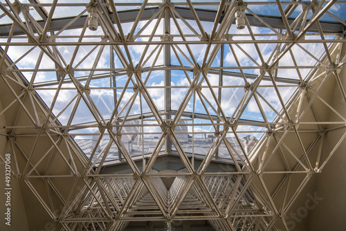Modern architectural fiction. A metal structure supporting contemporary gallery glass dome. Transparent ceiling letting in sunlight, glass dome blue sky and rooftop in background.