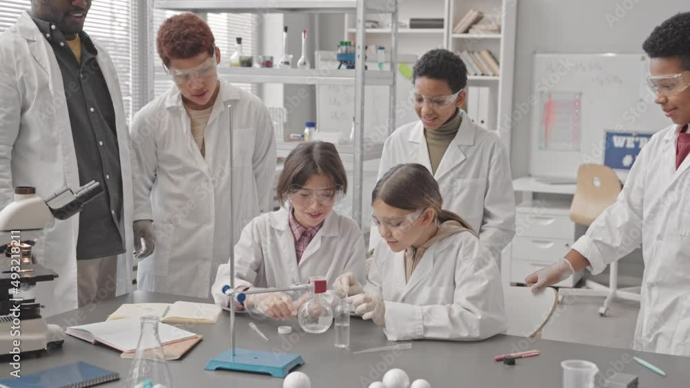 Slowmo shot of diverse school children and African American male teacher in lab coats doing chemical experiments at Chemistry class having fun together Slowmo shot of diverse school children and African American male teacher in lab coats doing chemical experiments at Chemistry class having fun together