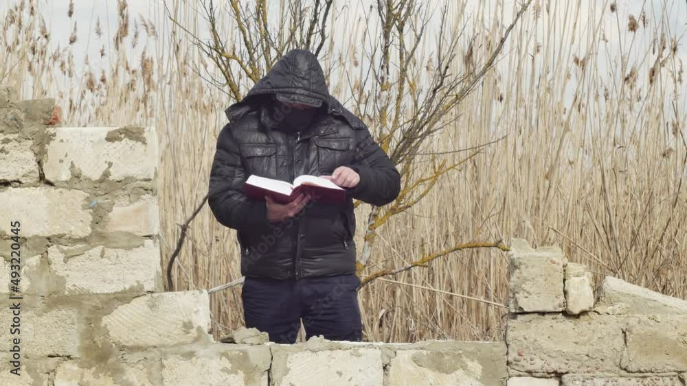 a man with a hood on his head reads a thick book standing behind a collapsed wall against the background of reeds a man with a hood on his head reads a thick book standing behind a collapsed wall against the background of reeds