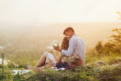 Young couple in love enjoying a glass of wine and a romantic picnic. The man gently hugs and kiss his young woman. 