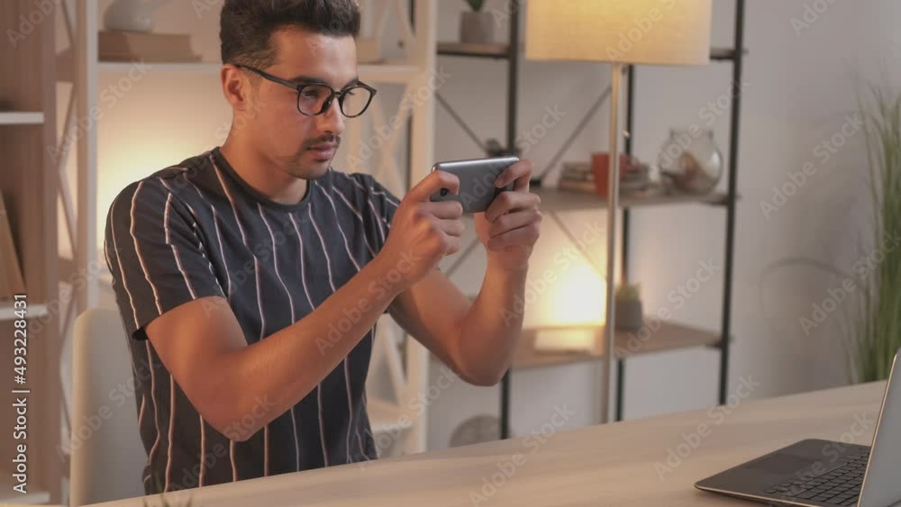 Mobile games. Addicted man. Virtual entertainment. Casual guy being involved in playing on smartphone sitting work desk with laptop light room interior. Mobile games. Addicted man. Virtual entertainment. Casual guy being involved in playing on smartphone sitting work desk with laptop light room interior.