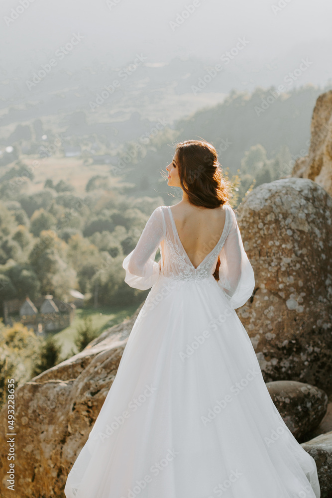 Gorceous blonde bride against the backdrop of the mountains in Italy. Bride in white dress standing on the hillside and looking at the sunset. on top of the mountain.