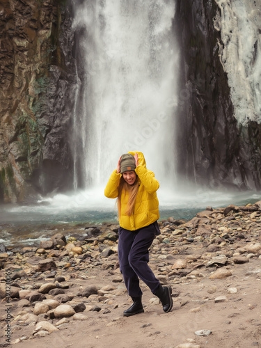 A woman stands crouched and laughing on the river bank against the backdrop of a stormy waterfall flowing from the ska. Optical illusion.