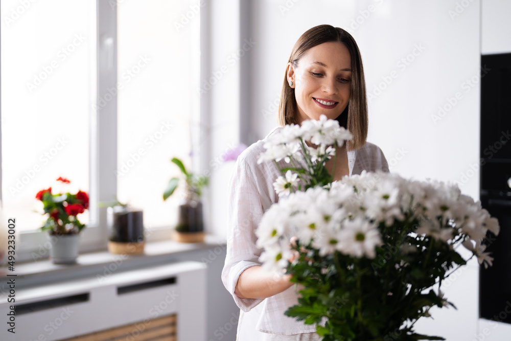 Happy and joyful young woman in white arranging white flowers at home in the kitchen Happy and joyful young woman in white arranging white flowers at home in the kitchen