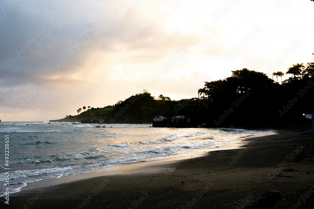 Sunrise over tropical beach, mountain and palm trees. Beautiful view in Dominican Republic. Cloudy sky and golden rays of sun. 