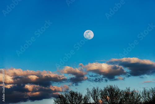 An almost full moon is rising above the trees and clouds on this early evening in March in Upstate NY.