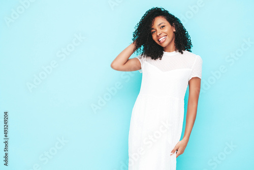 Beautiful black woman with afro curls hairstyle. Smiling model dressed in white summer dress. Sexy carefree female posing near blue wall in studio. Tanned and cheerful. Isolated © halayalex