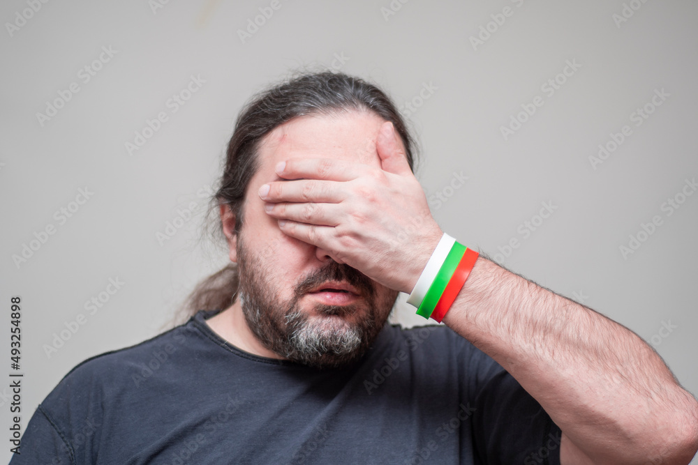 Fototapeta premium An unshaven man with bracelets in the colors of the Bulgarian flag pathetically closes his eyes with his palm.