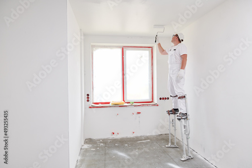 Painter worker on stilts with roller painting ceiling into white photo