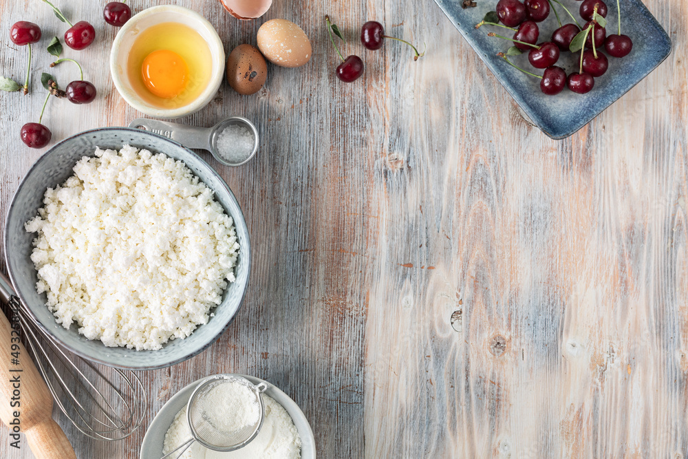 Cottage cheese, egg, flour and fresh cherries are the ingredients for a cottage cheese casserole on a wooden table.