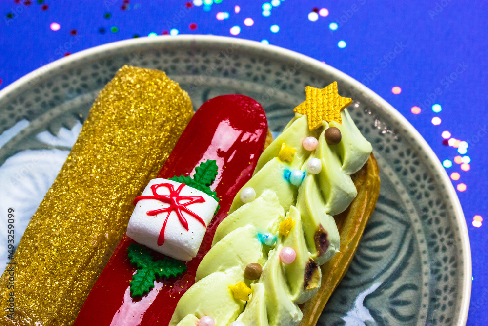 Festively decorated gold, green, red eclairs in New Year's style 2022 lying on round gray plate top view. Eclairs decorated with Christmas tree, gift box, sequins. Blue table with colorful confetti.