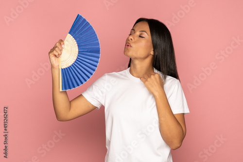Exhausted african american woman with closed eyes using blue fan suffer from heat sweating, feels sluggish. Black female cooling in hot summer weather, high temperature, isolated on pink background. photo