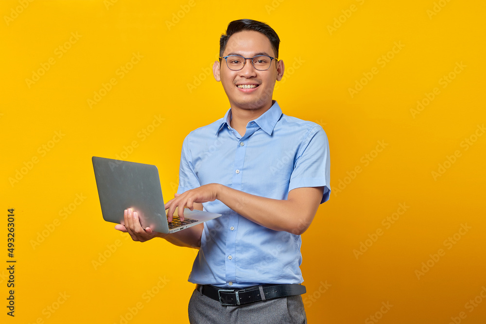 Portrait of cheerful handsome Asian man in glasses using laptop isolated on yellow background. businessman and entrepreneur concept Portrait of cheerful handsome Asian man in glasses using laptop isolated on yellow background. businessman and entrepreneur concept
