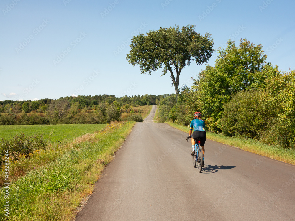 a single woman from the back, doing gravel bike on a quiet countryside road, on a summer sunny day