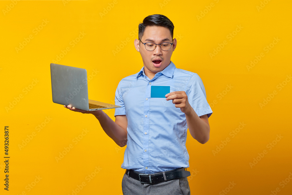 Surprised young asian man Asian in glasses holding laptop and looking credit card isolated on yellow background. businessman and entrepreneur concept Surprised young asian man Asian in glasses holding laptop and looking credit card isolated on yellow background. businessman and entrepreneur concept