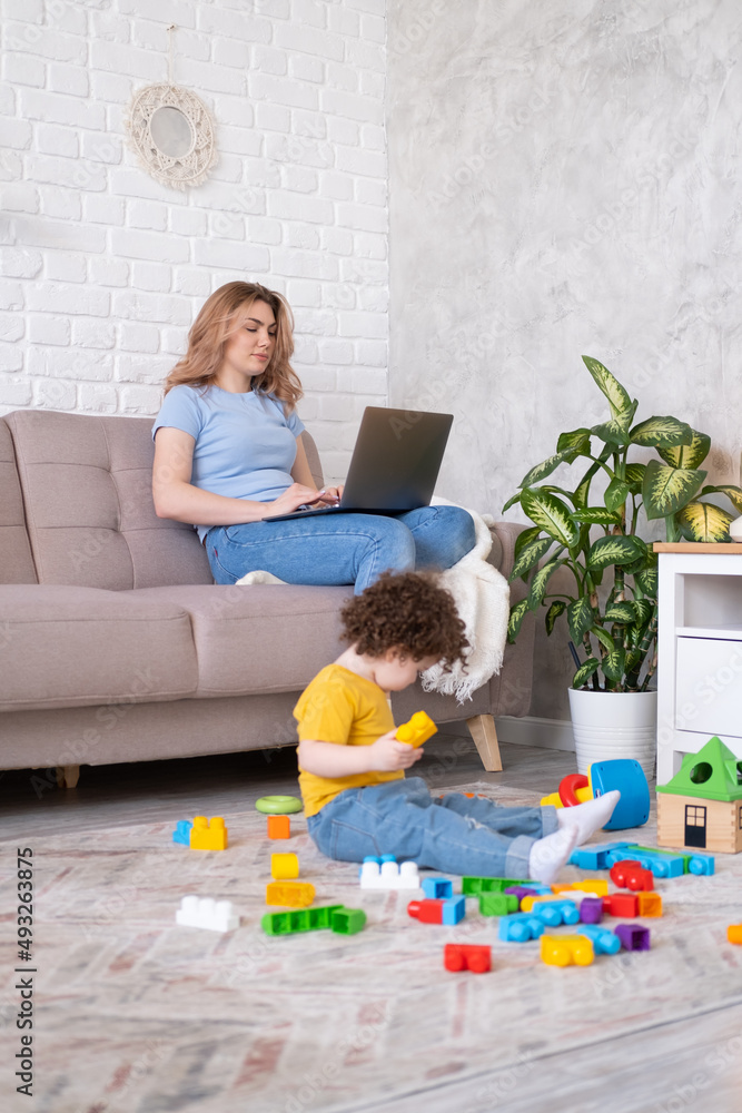 Mother freelancer use laptop sits on couch, kid daughter plays with toys on floor. Working from home Mother freelancer use laptop sits on couch, kid daughter plays with toys on floor. Working from home
