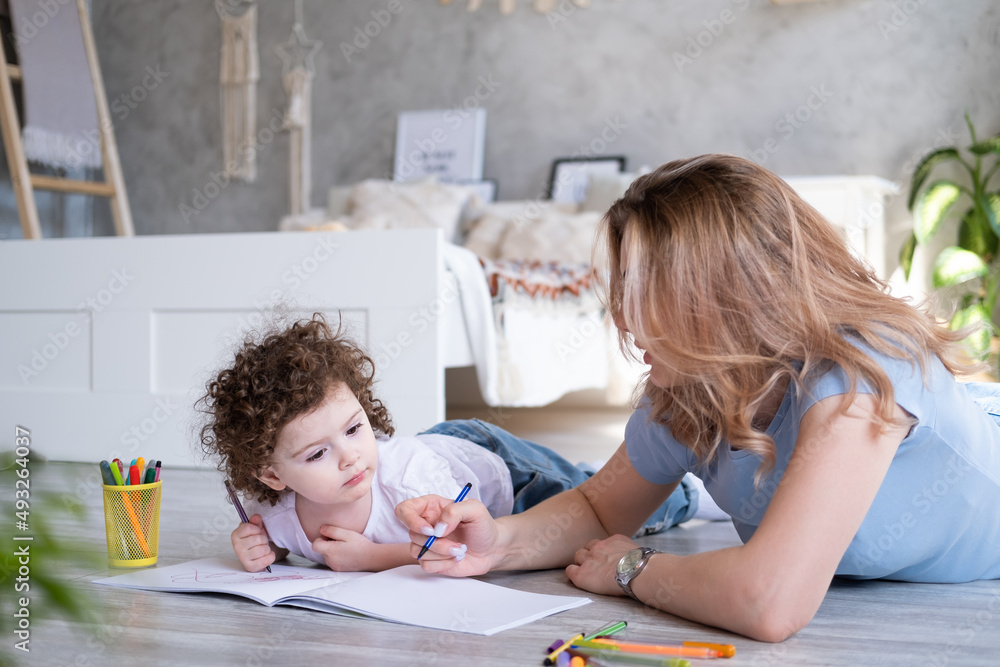 young mother and curly child girl drawing with colored markers lying on floor room at home