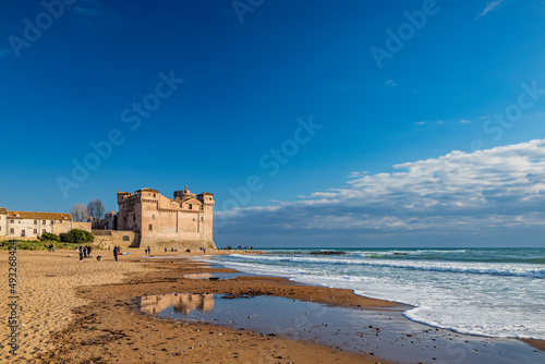Santa Marinella, Rome, Lazio, Italy - The seaside castle of Santa Severa on a cold winter day. The rough sea, the strong wind, the cloudy blue sky. Many people stroll and enjoy the sun on the beach. © Ragemax