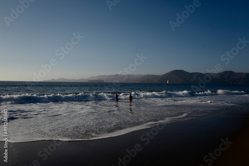 Ocean water as waves with human children swimming and sailboat in distance.
