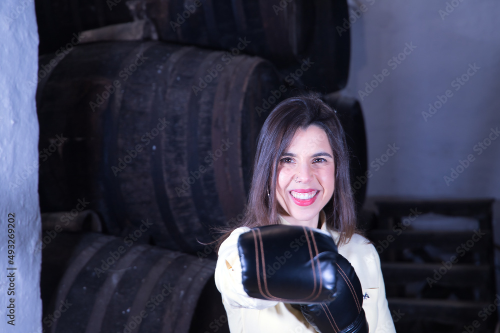 Portrait of young, beautiful and feisty woman in yellow shirt and jeans, with boxing gloves in her hands next to the barrels in her cellar. Concept businesswoman, entrepreneur, drink, wine, alcohol.