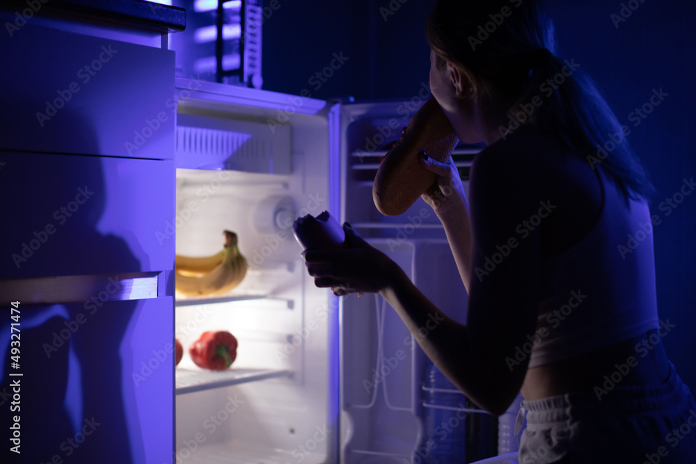 A woman eats bread and sausage while sitting near an open refrigerator in the middle of the night.