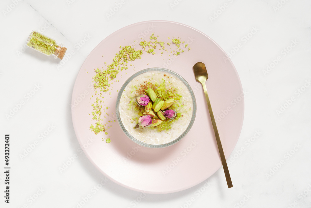 Creamy rice pudding topped with pistachio and cinnamon in a glass bowl on pink plate and marble table. Traditional Turkish dessert sutlac. Minimal bright and airy background.  Copy space. Top view. 