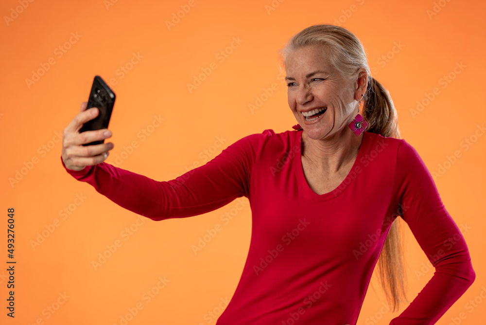 Pretty mature woman using mobile cell phone doing selfie greet with hand isolated on solid orange background studio portrait Pretty mature woman using mobile cell phone doing selfie greet with hand isolated on solid orange background studio portrait