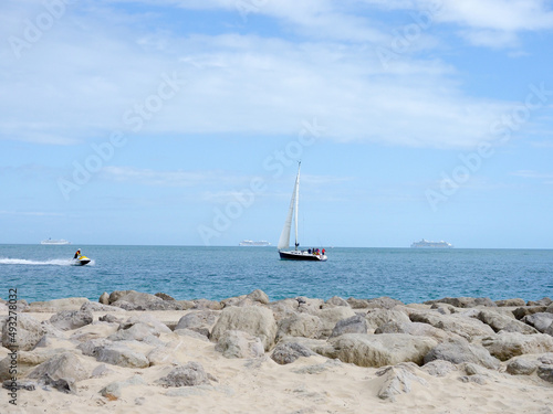 A sailing yacht passing close to the shore with three anchored cruise liners in the distance and a jet ski passing by photo