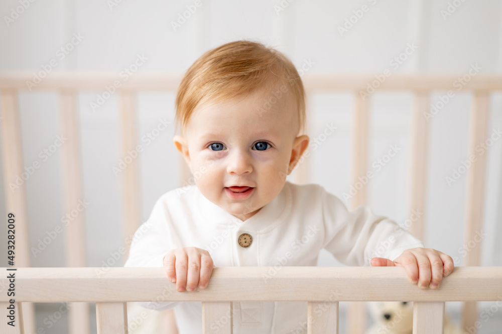 portrait of a 6-month-old blond boy standing in a crib in a bright children's room and smiling in a cotton bodysuit, the concept of children's goods portrait of a 6-month-old blond boy standing in a crib in a bright children's room and smiling in a cotton bodysuit, the concept of children's goods